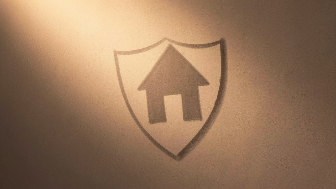 A shield-like symbol cast in soft shadow on a wall, with a house silhouette integrated, conveying protection.